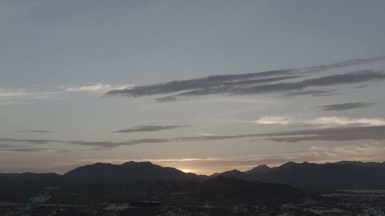 Wide Los Cabos, Mexico sunset view with dark mountain ridges, layered clouds, a thin warm glow near the horizon, and no visible people.