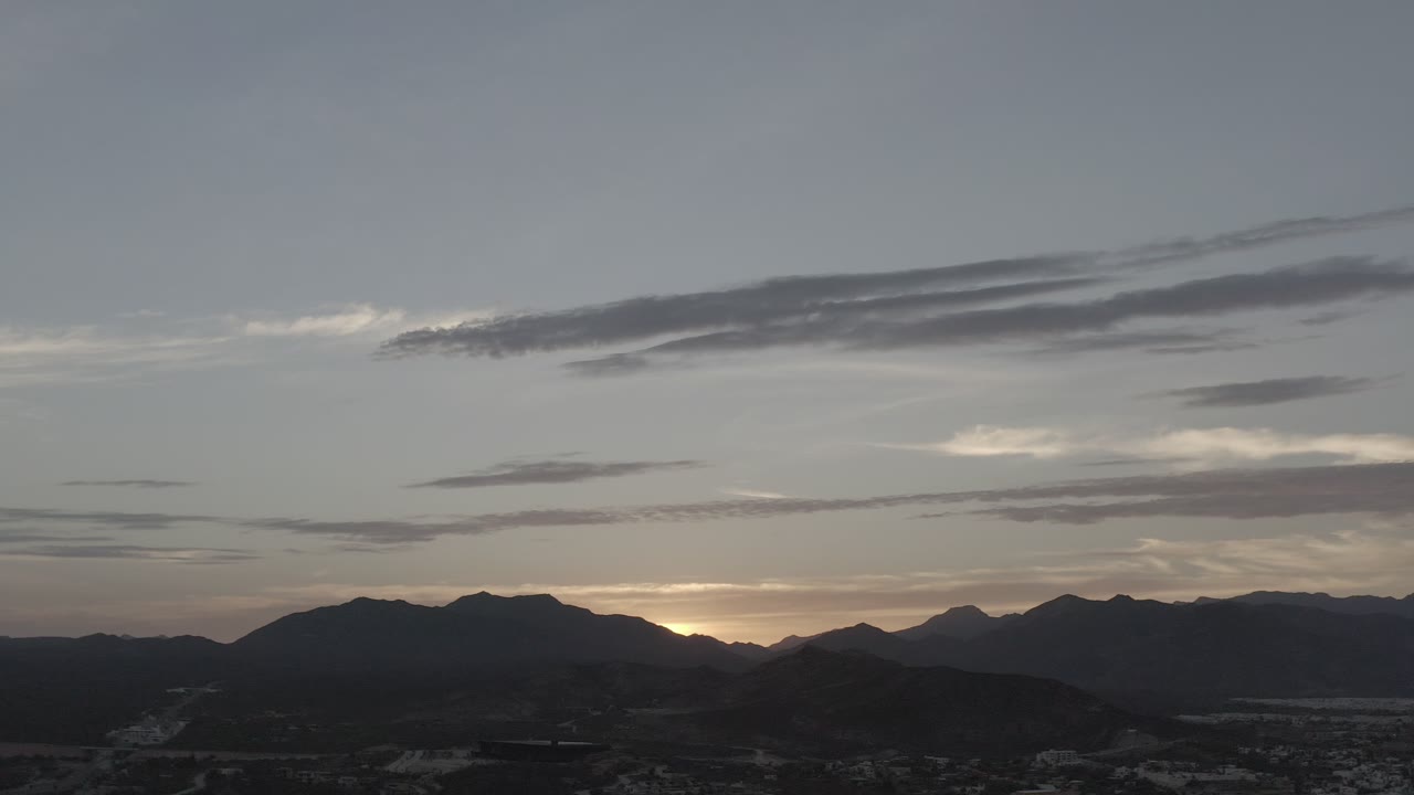 Los Cabos mountain ridges sit in silhouette below broad gray-blue sky, with long cloud streaks and a small warm opening at the horizon, no visible people.