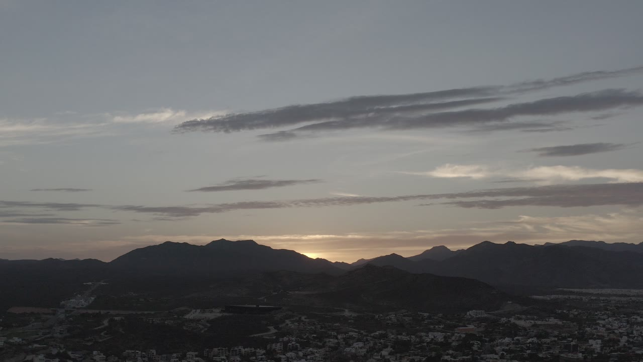 Opening frame of Los Cabos, Mexico at dawn, with dark mountain ridges, a faint sunrise glow behind the horizon, a gray cloudy sky, and no visible people.