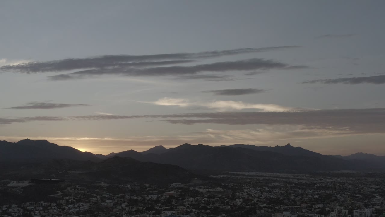 Opening view of Los Cabos at dusk with dark mountain ridges, layered clouds, town buildings below, and no visible people.