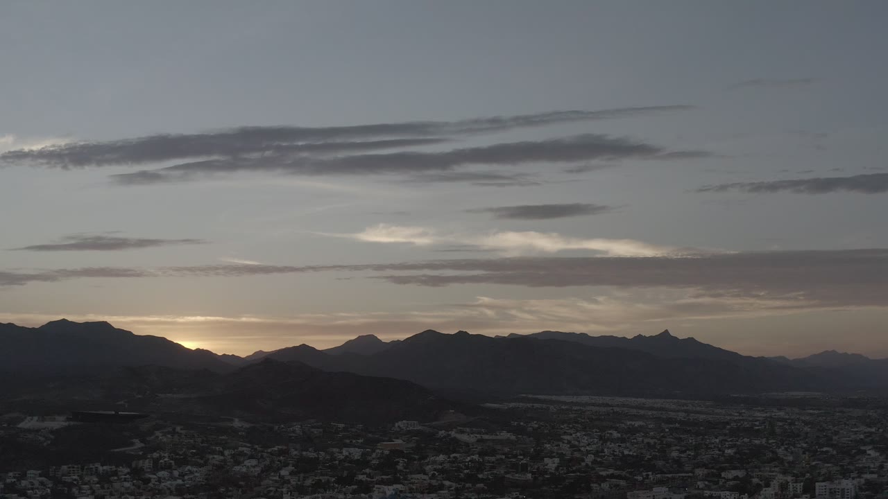 Early frame showing Los Cabos town in shadow below a broad mountain skyline and thin horizontal clouds, with no visible people.