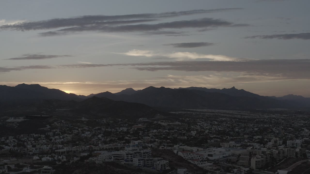 Wide elevated view in Los Cabos, Mexico, showing a dark community foreground, layered mountain silhouettes, gray clouds, a thin sunrise glow on the horizon, and no visible people.