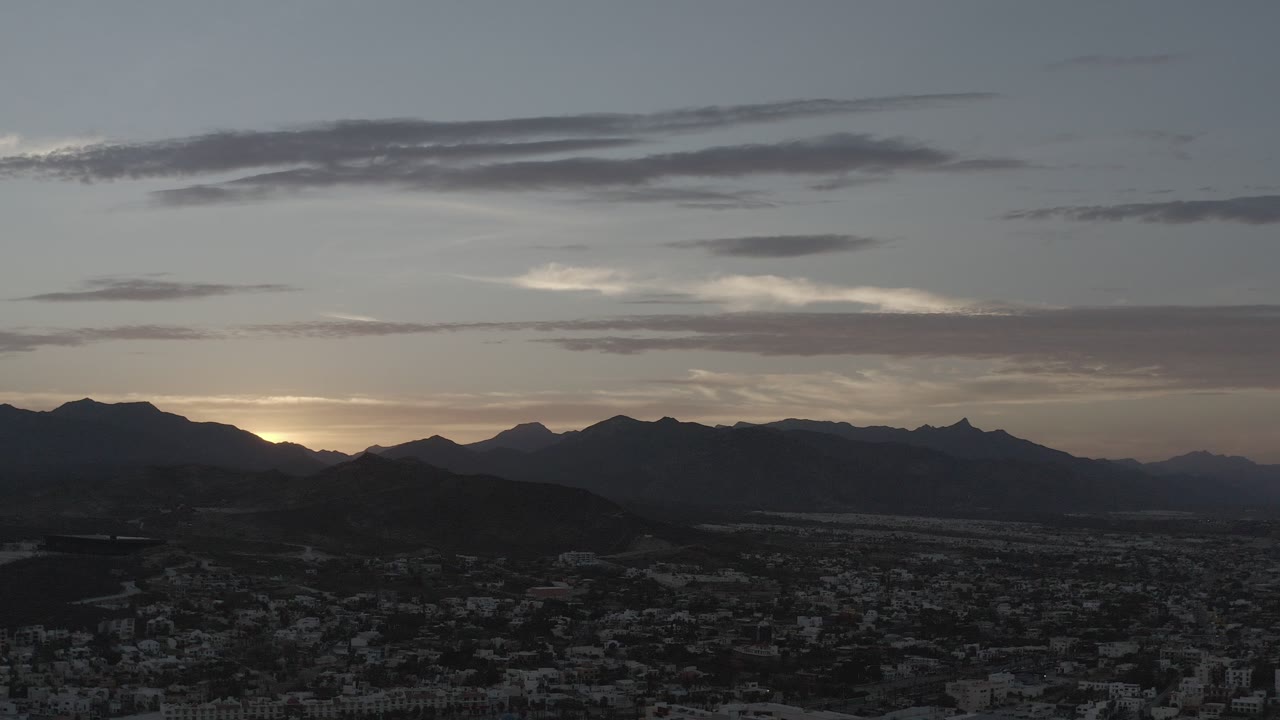 Los Cabos, Mexico sunrise begins behind a dark mountain ridge, with a low city foreground, gray-blue clouds, and no visible people.