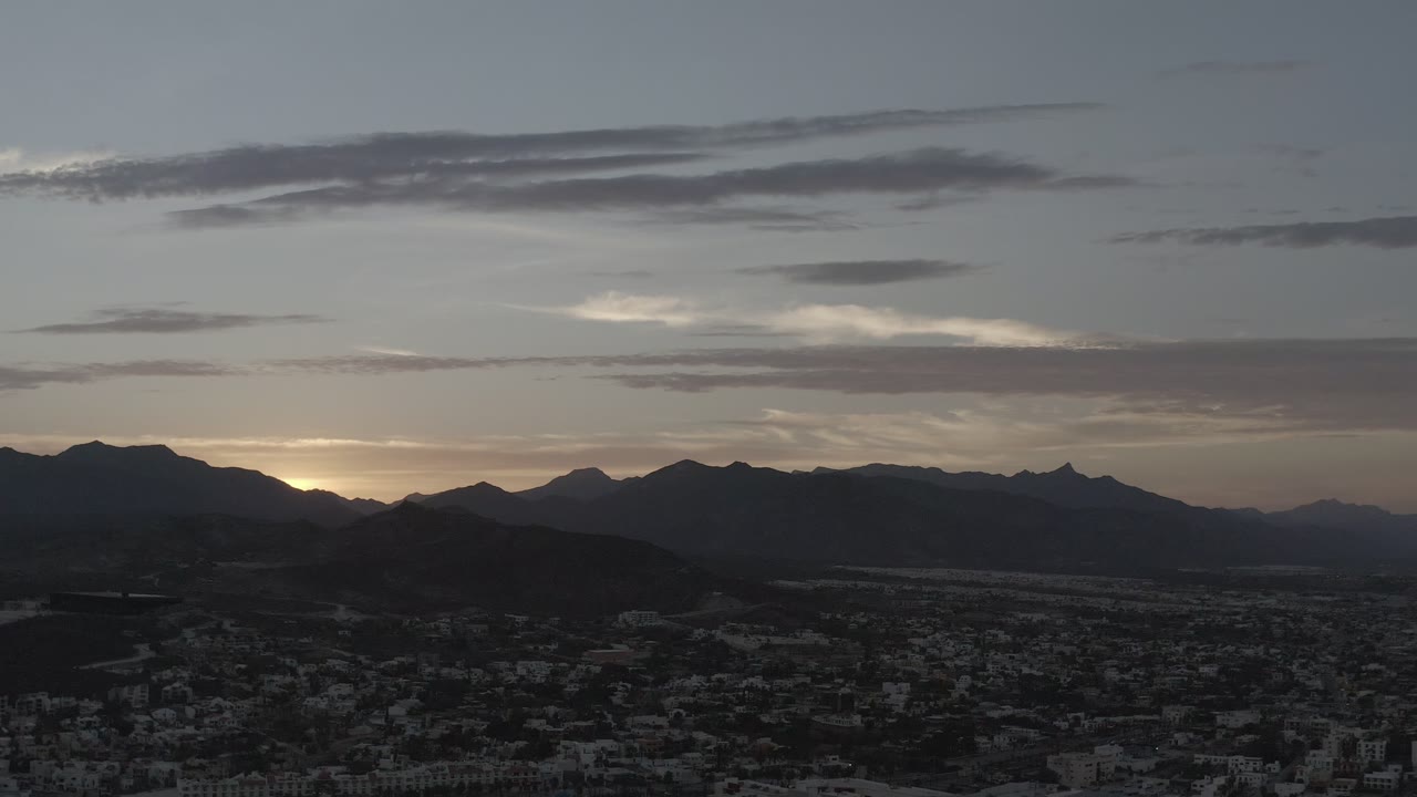 A wide Los Cabos dawn view shows layered black mountain shapes, scattered clouds, a pale horizon glow, and no visible people.