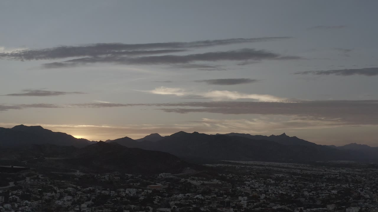 Los Cabos mountain silhouettes stretch across the frame beneath a gray cloud bank, with the town still visible in shadow and no visible people.