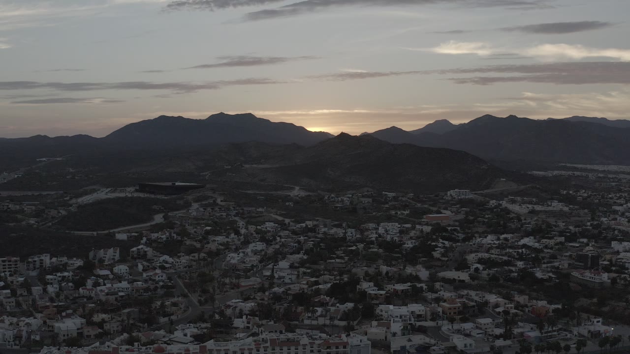 Opening frame of Los Cabos, Mexico, showing silhouetted mountain ridges, a dim valley with buildings below, pale clouds, and no visible people.