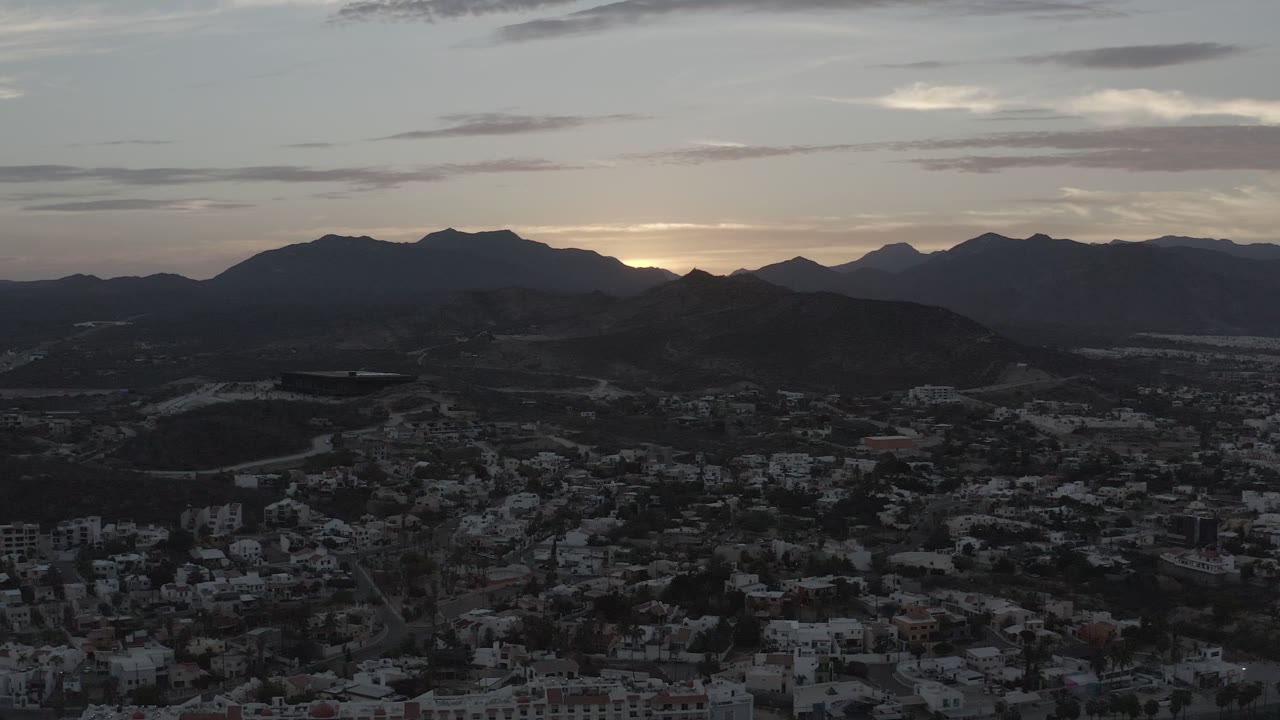 Early frame of Los Cabos, Mexico, with a dark central ridge, clustered buildings in the lower valley, thin clouds overhead, and no visible people.