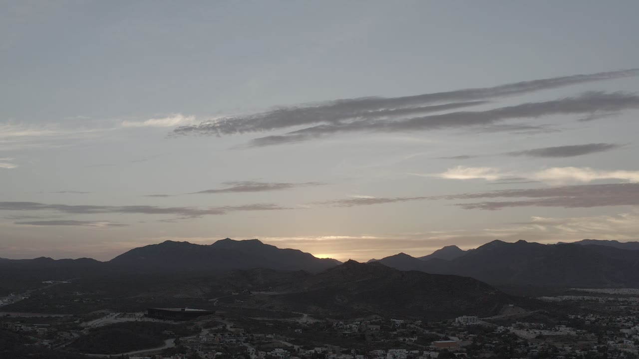 Opening frame of Los Cabos at dusk, with dark mountain silhouettes, a low band of town detail, thin clouds, and no visible people.