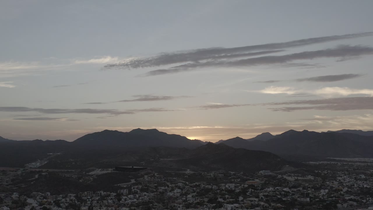 Early frame showing the Los Cabos mountain horizon under long gray cloud bands, with the town remaining dark at the bottom and no visible people.
