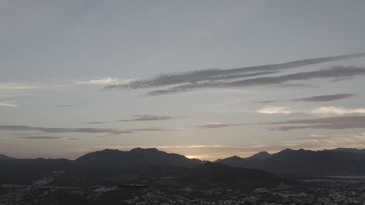 Los Cabos, Mexico sunset view with dark mountain silhouettes, a faint city foreground, layered clouds, and no visible people.