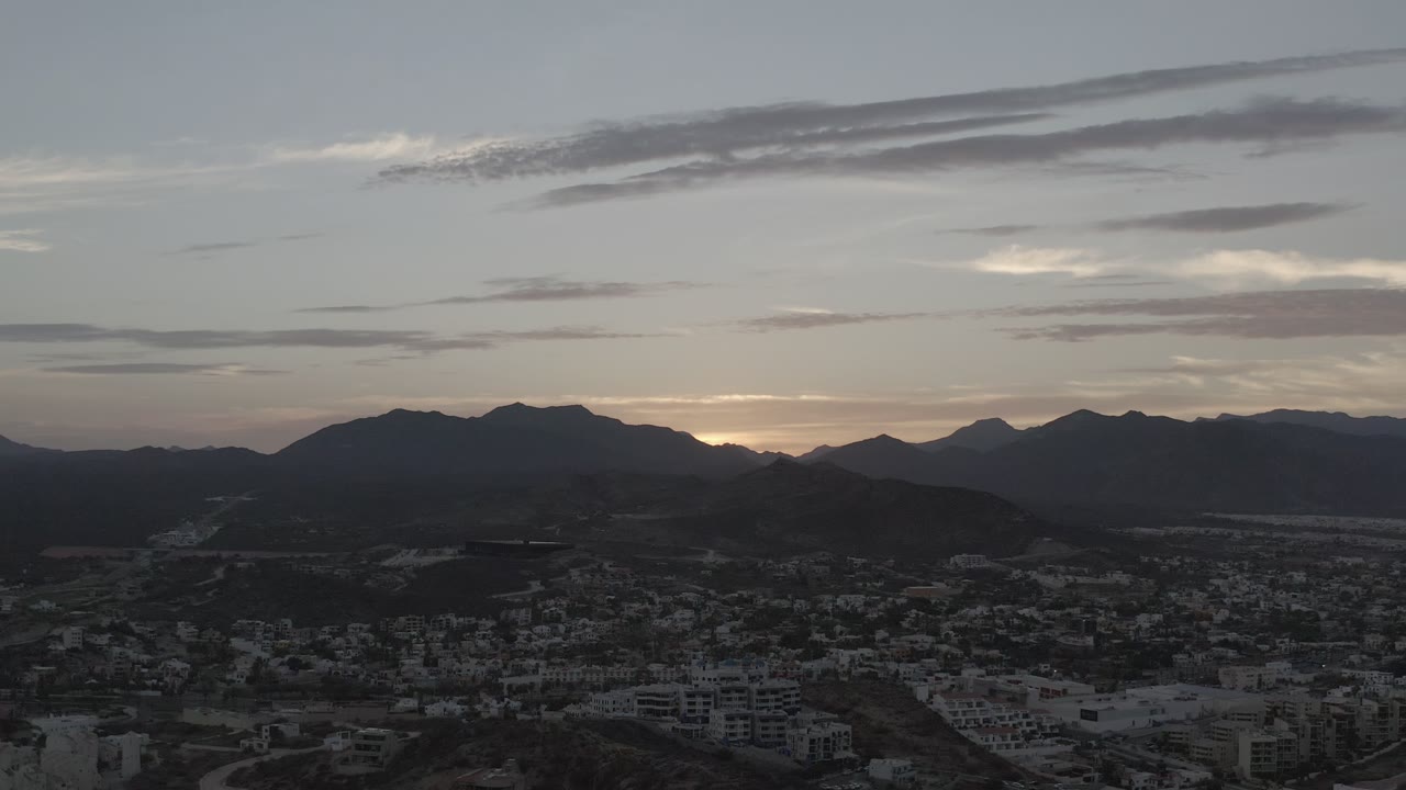 Los Cabos townscape remains dark in the foreground while a thin band of early sunrise light appears behind the mountain ridge and stretched cloud layers, with no visible people.