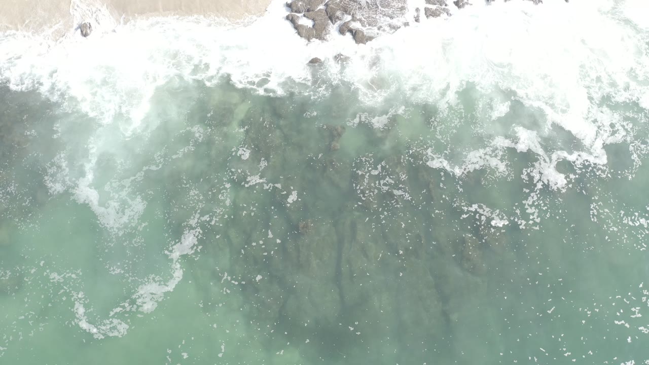 Overhead view in Los Cabos, Mexico of white surf washing along a bright sandy edge above pale turquoise water, with no visible people.