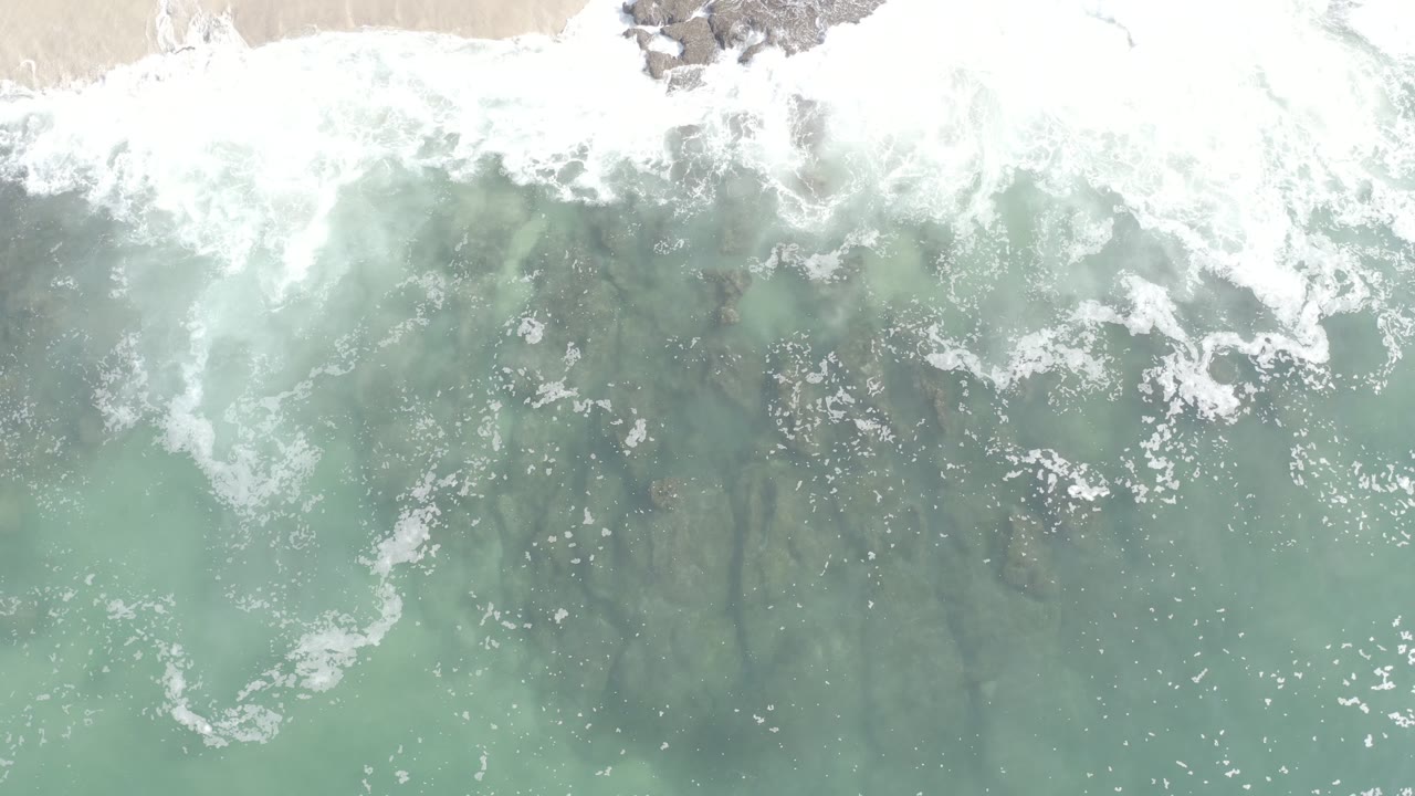 Aerial view of foamy wave water with dark rocks under the surface.