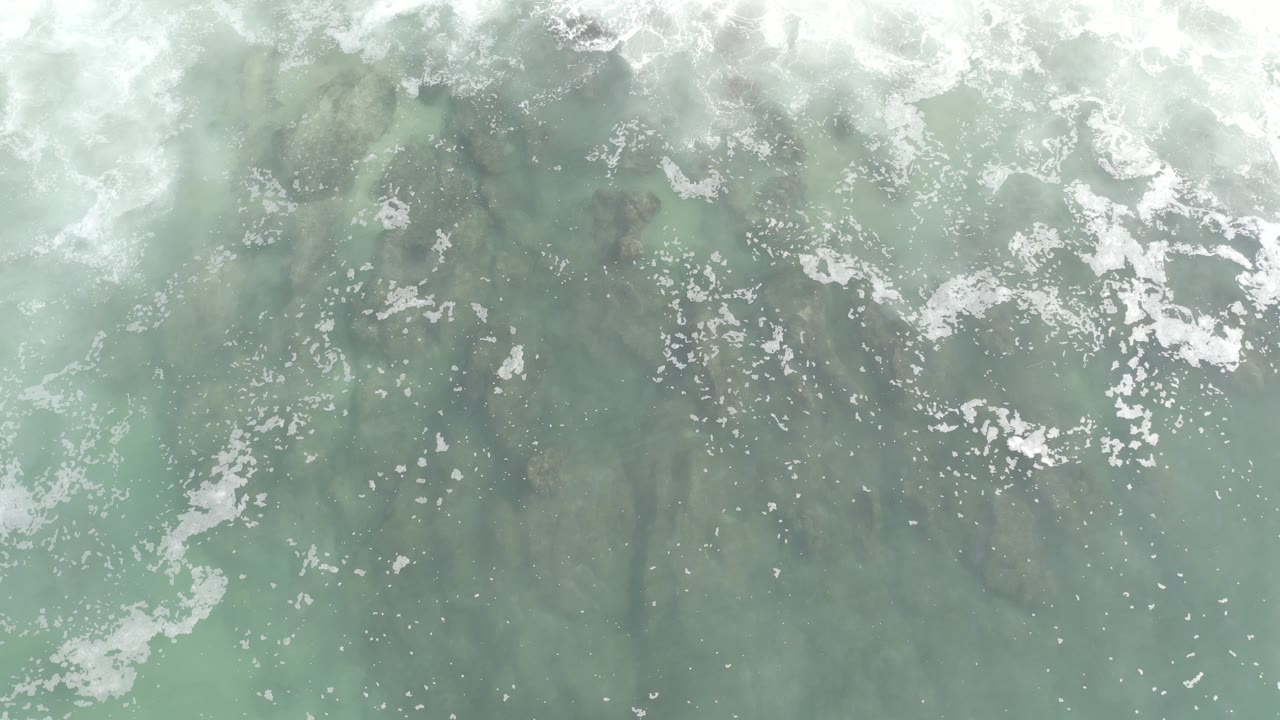 Overhead view of pale green water in Los Cabos with white foam scattered near the top edge and no visible people.