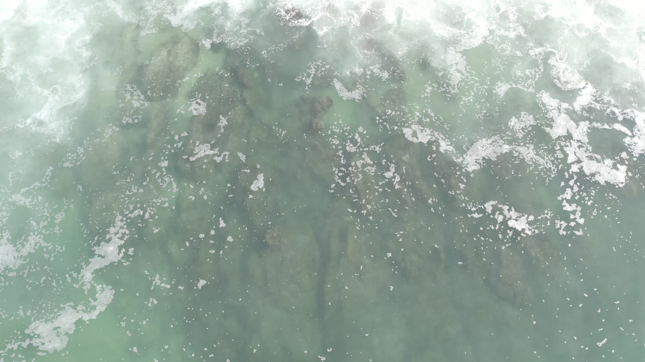 Overhead Los Cabos water frame with clustered white bubbles across the upper half and muted green-blue water below, with no visible people.