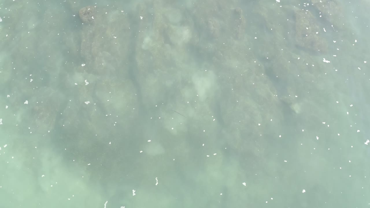 Pale gray-blue water fills the Los Cabos frame from above, with subtle ripples, murky streaks beneath the surface, scattered foam, and no visible people.