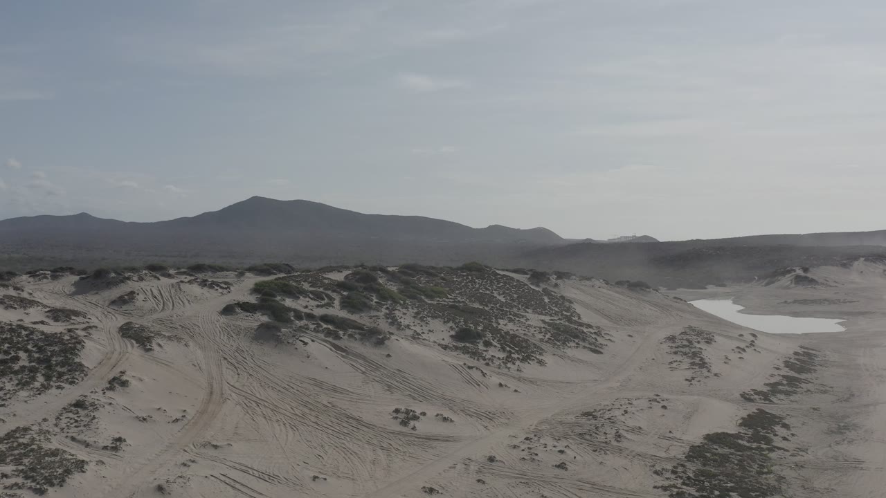 Wide aerial view of Los Cabos desert sand crossed by tire tracks, low shrubs, hazy sky, distant mountains, and no visible people.