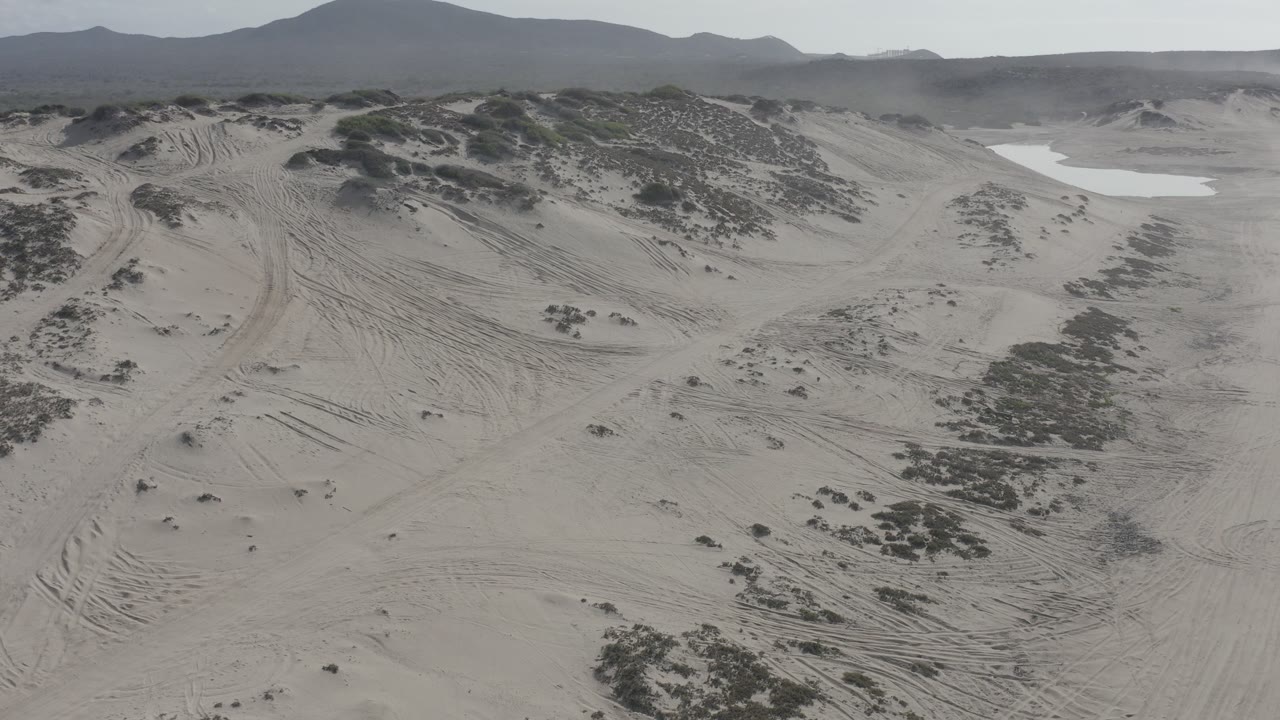 Aerial view over Los Cabos sand dunes with curved tire tracks, dark scrub patches, distant hills, muted daylight, and no visible people.