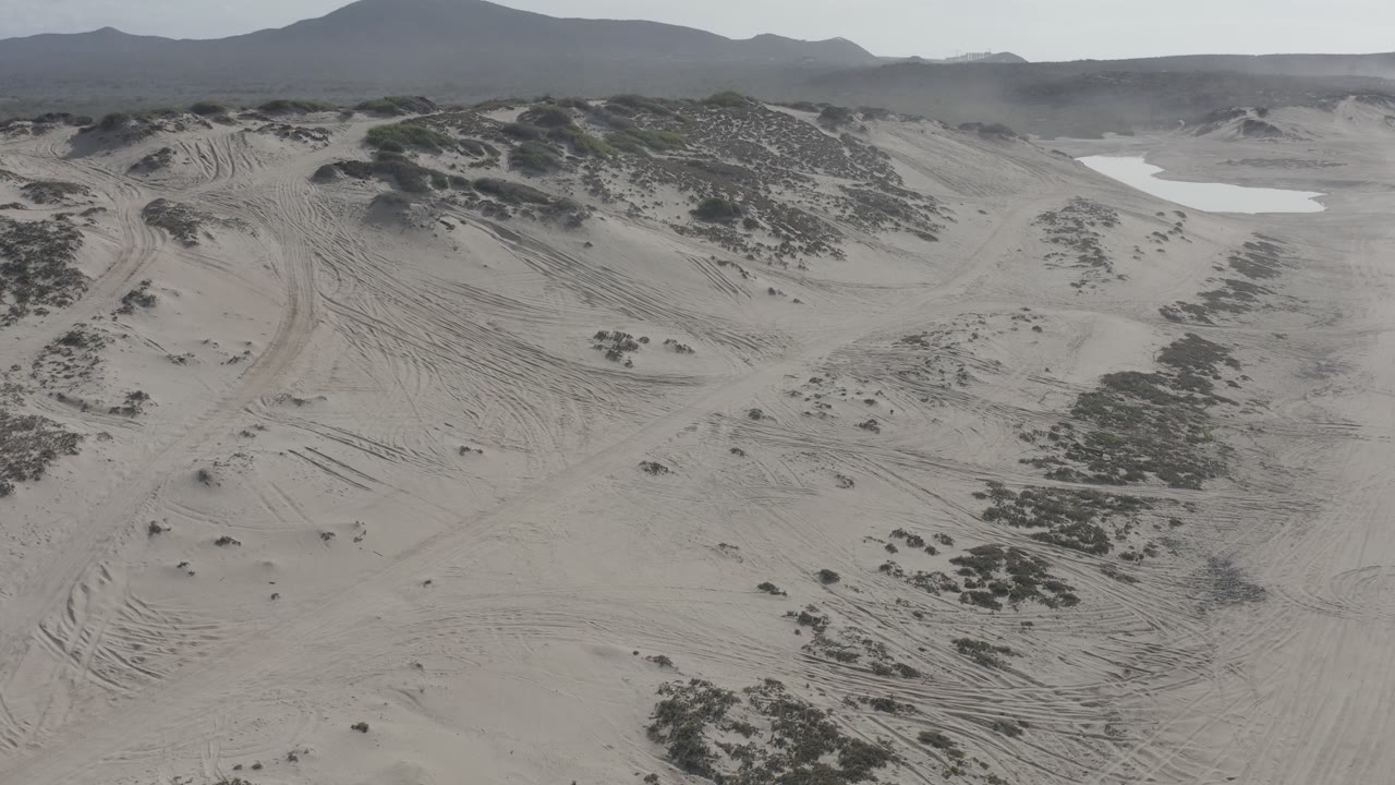Los Cabos aerial dune view with pale sand, low scrub, visible vehicle tracks, a bright sandy basin near the horizon, and no visible people.
