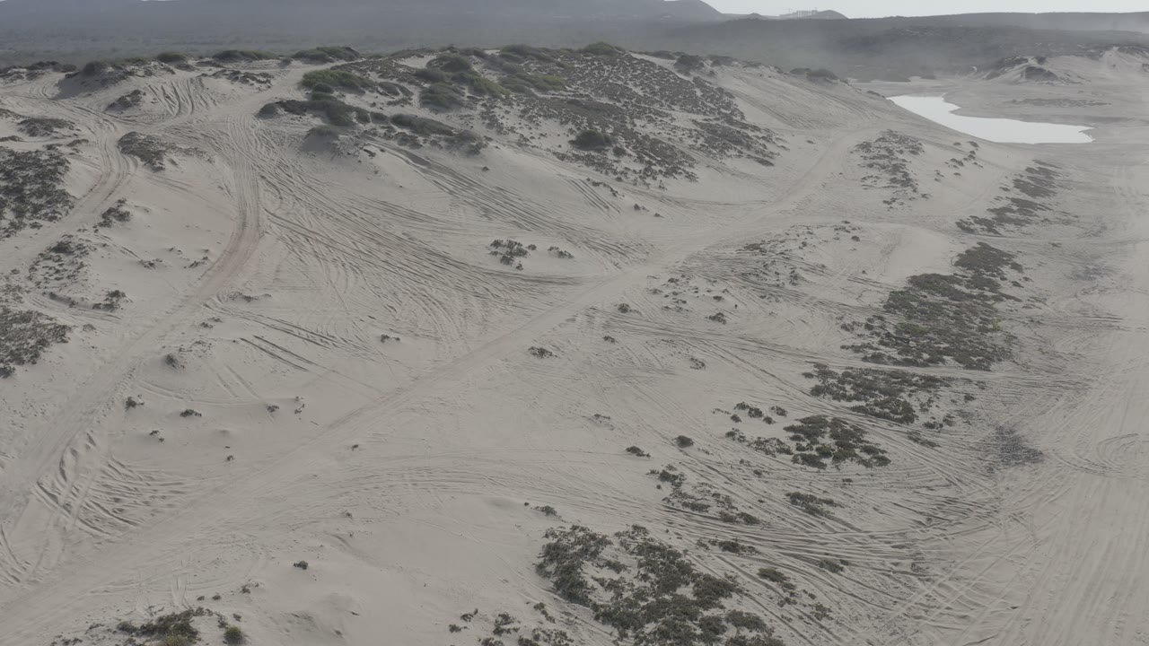 Wide aerial view of rutted sand dunes with scrub clusters and hills on the horizon.