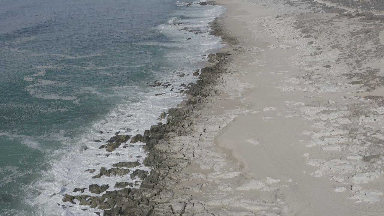 Aerial view in Los Cabos, Mexico, with blue-green ocean at left, white surf on dark rocks, pale sand at right, and no visible people.