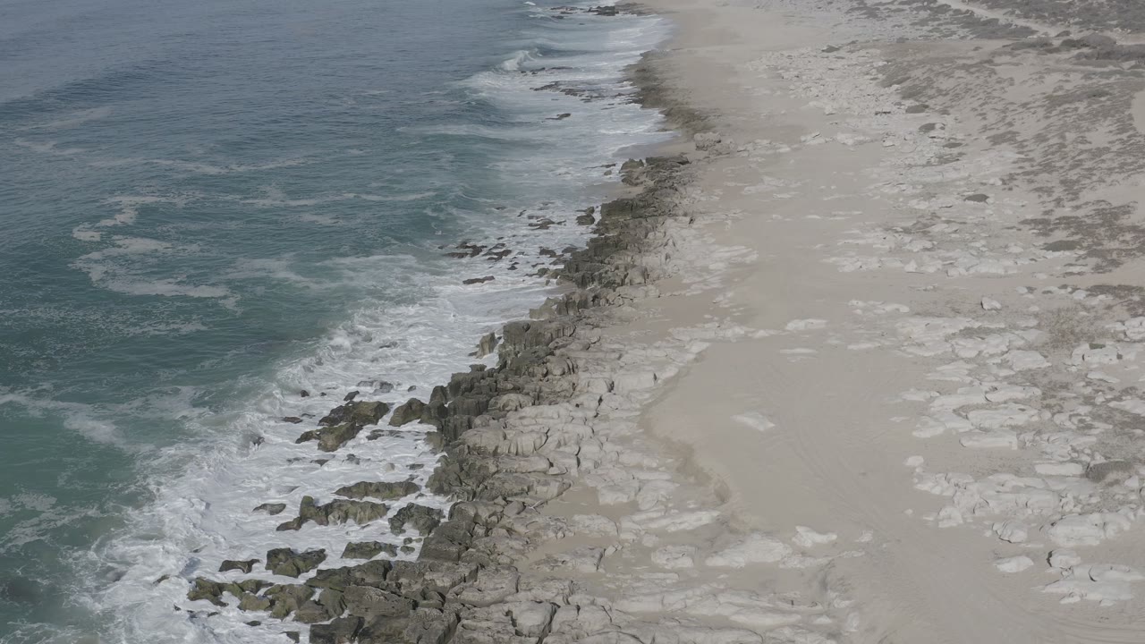 Los Cabos shoreline from above, with a thicker band of white water curling against the rocks, open sand to the right, and no visible people.