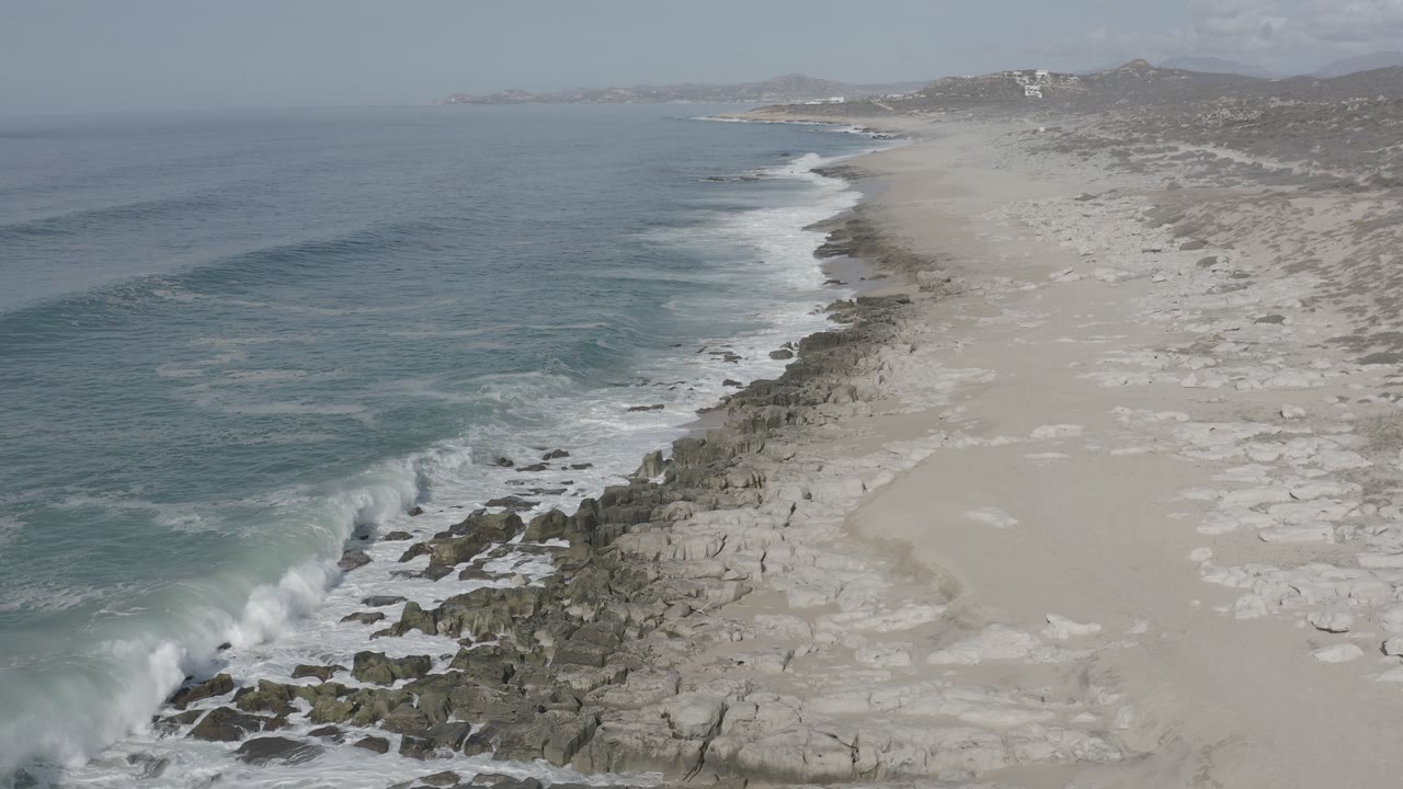 Elevated view of the Los Cabos shoreline with blue-gray ocean on the left, pale beach on the right, rocks down the center, and no visible people.