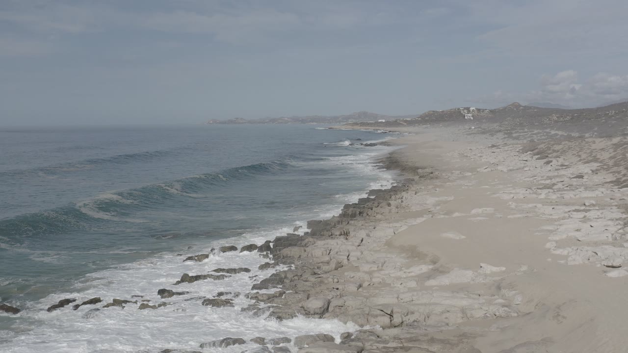 Elevated view of the Los Cabos shoreline with blue-gray ocean on the left, pale sand and rocks on the right, and no visible people.