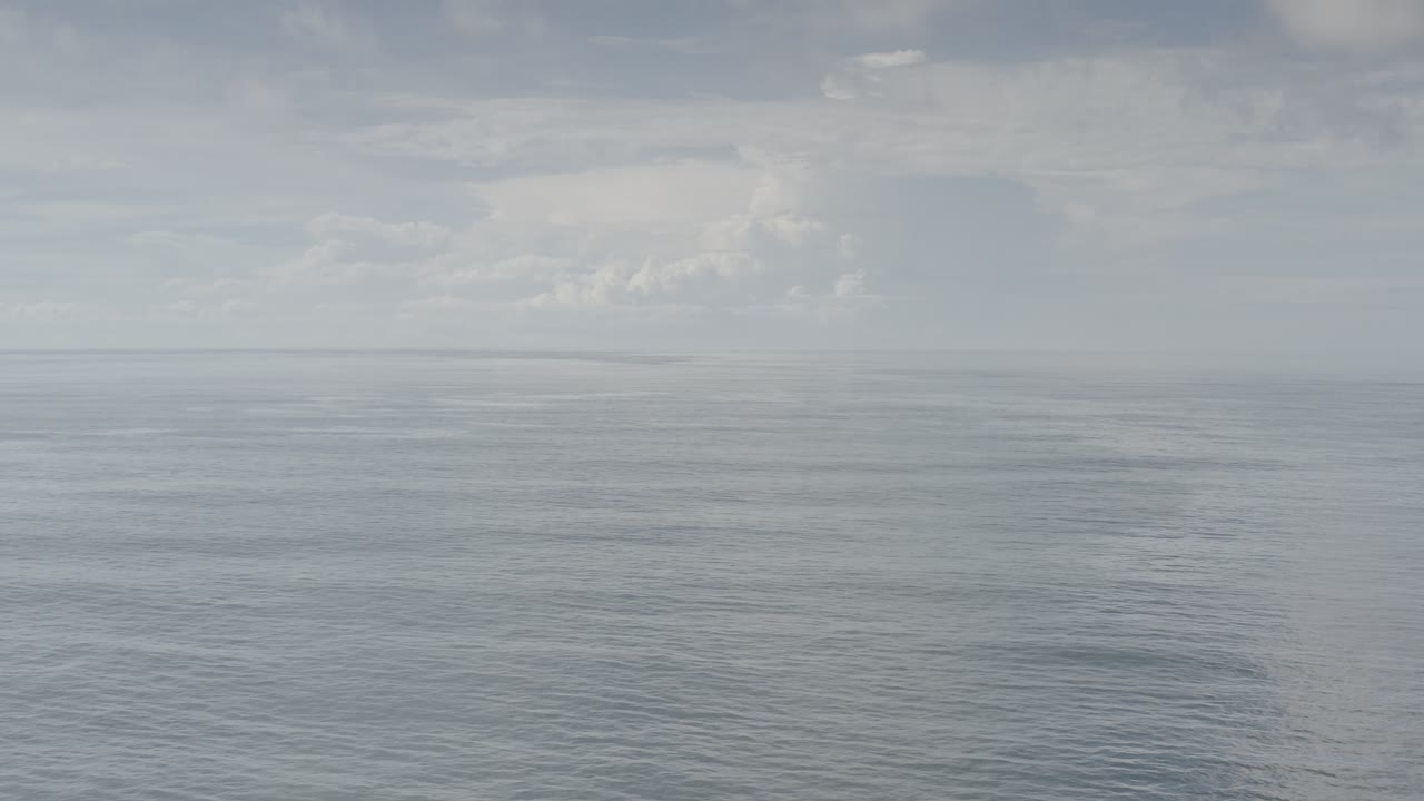Opening frame of Los Cabos ocean from above, with soft cloud cover, a flat horizon, faint distant land, and no visible people.