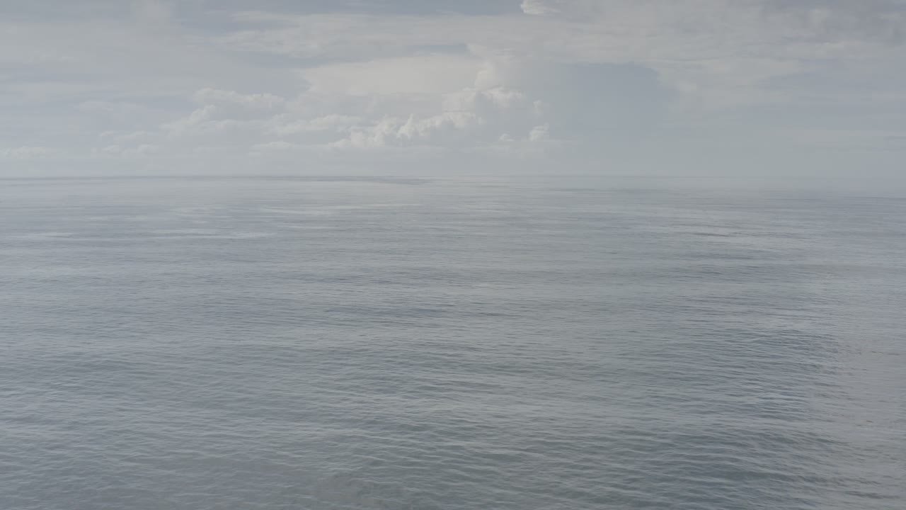 Opening frame of calm blue-gray ocean and cloudy haze in Los Cabos, Mexico, with a faint distant land shape at left and no visible people.