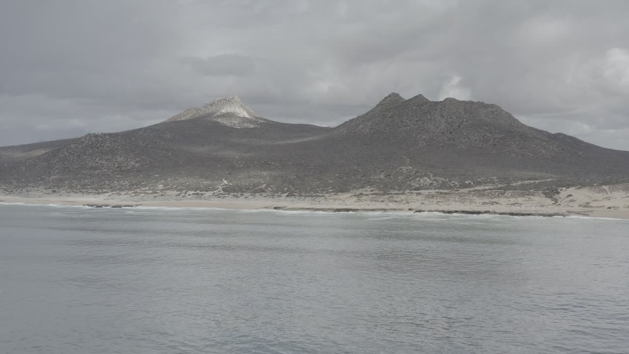 Opening view of Los Cabos, Mexico, with gray water in the foreground, a thin surf line on the beach, dark coastal mountains beyond, and no visible people.