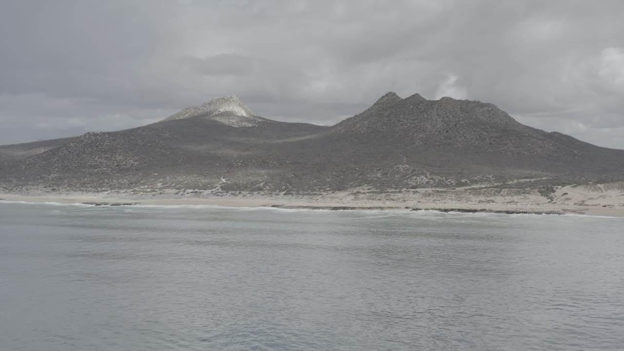 Wide beach and mountain range seen across calm gray water.