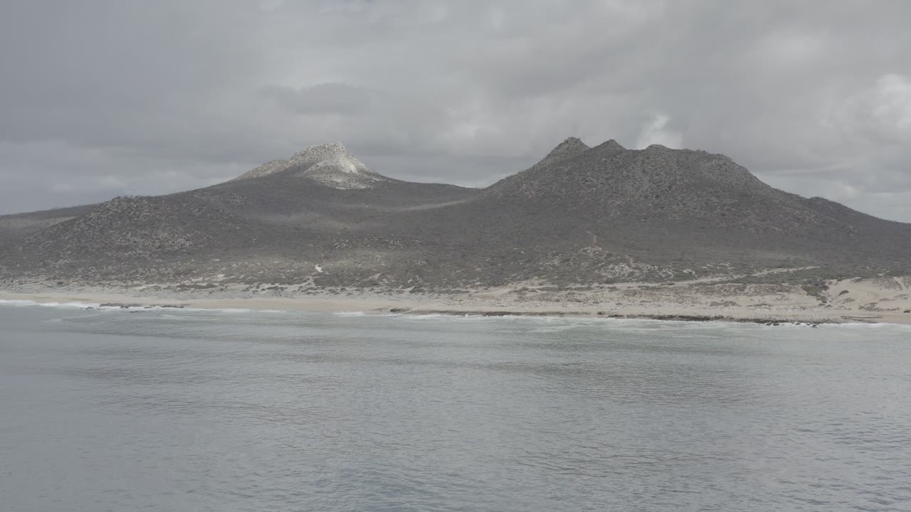 Wide coastal view of gray water, sandy beach, and two dark mountains under clouds.