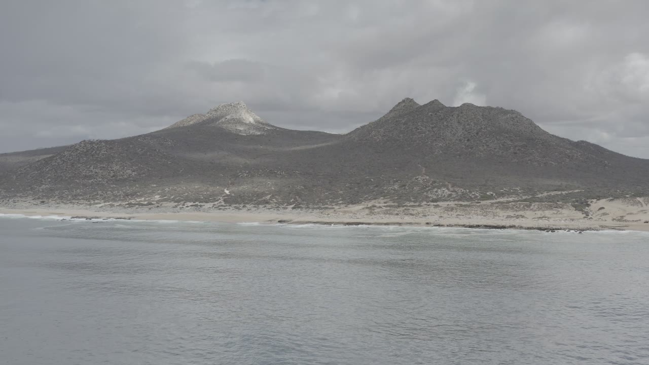 A wide Los Cabos beach view shows calm gray water, a narrow sand band, dark hills and mountains, and no visible people.