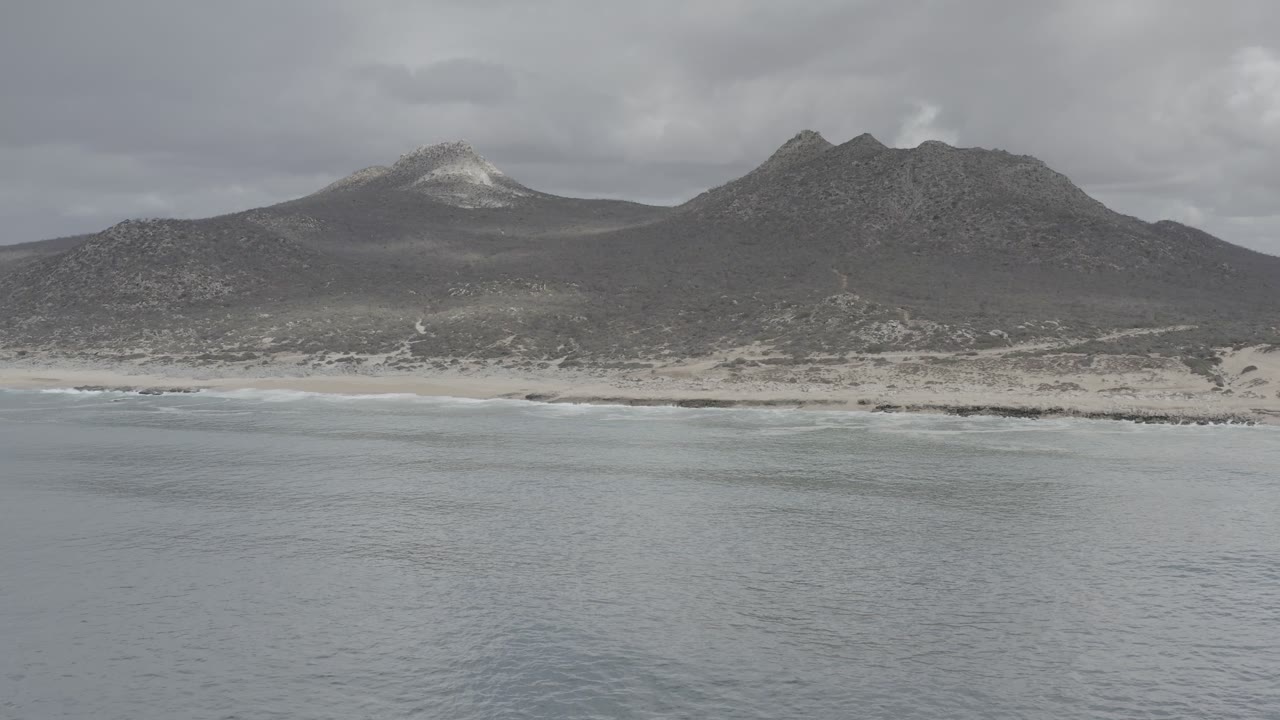 Second frame of the Los Cabos coast showing low surf against the beach, pale dunes, dark mountain ridges, clouded sky, and no visible people.