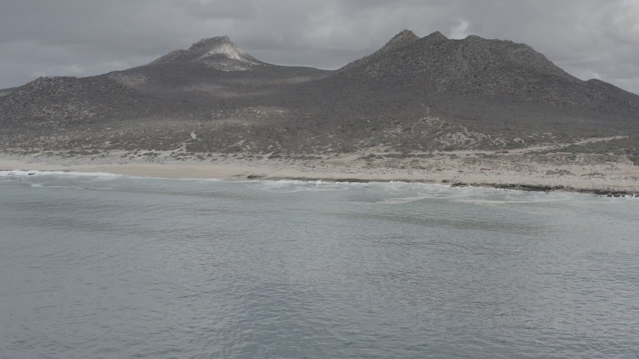 Wide aerial view of ocean, sandy beach, mountains, and cloudy sky.
