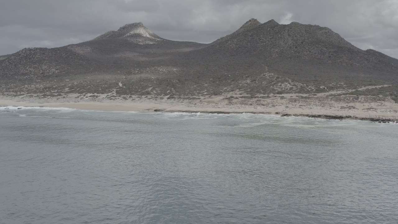Los Cabos shoreline seen from above, where the pale ocean reaches a sandy strip below dark mountain ridges and a gray cloud ceiling, with no visible people.