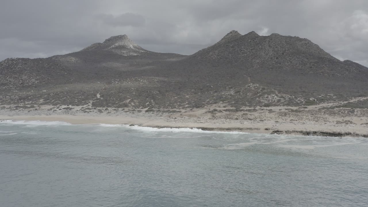 Wide aerial view of pale water, sandy beach, and dark hills under gray clouds.