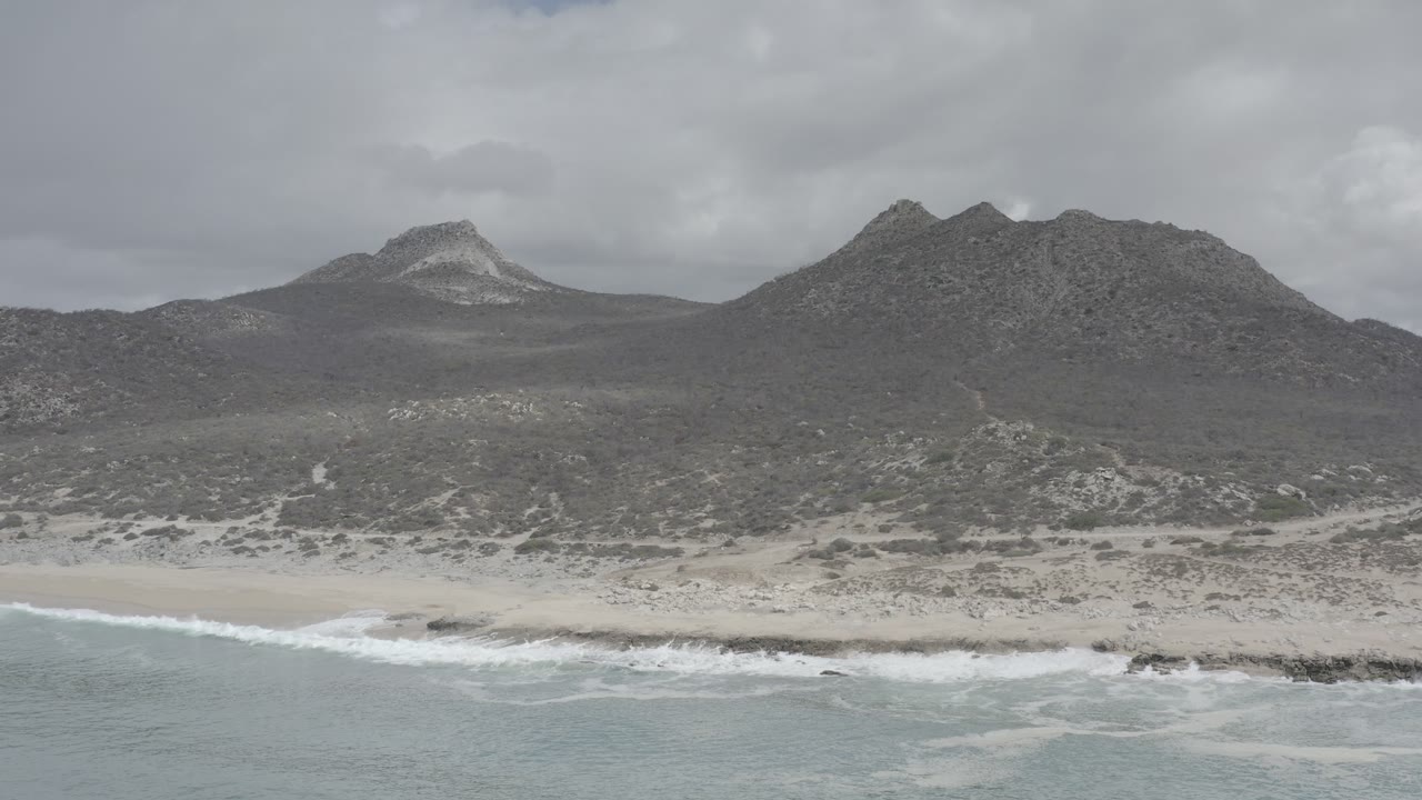 Aerial view of surf, sandy beach, scrubland, and rocky hills under gray clouds.