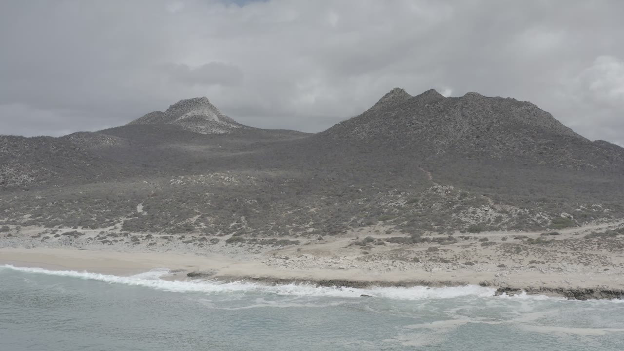 Aerial Los Cabos coastline view one second in, with a thin line of breaking waves, pale sand, scrubby terrain, layered rocky hills, and no visible people.