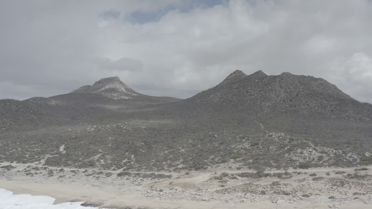 Los Cabos landscape with a rounded mountain ridge, scrub-covered flats, pale beach sand, and soft cloud cover, no visible people.