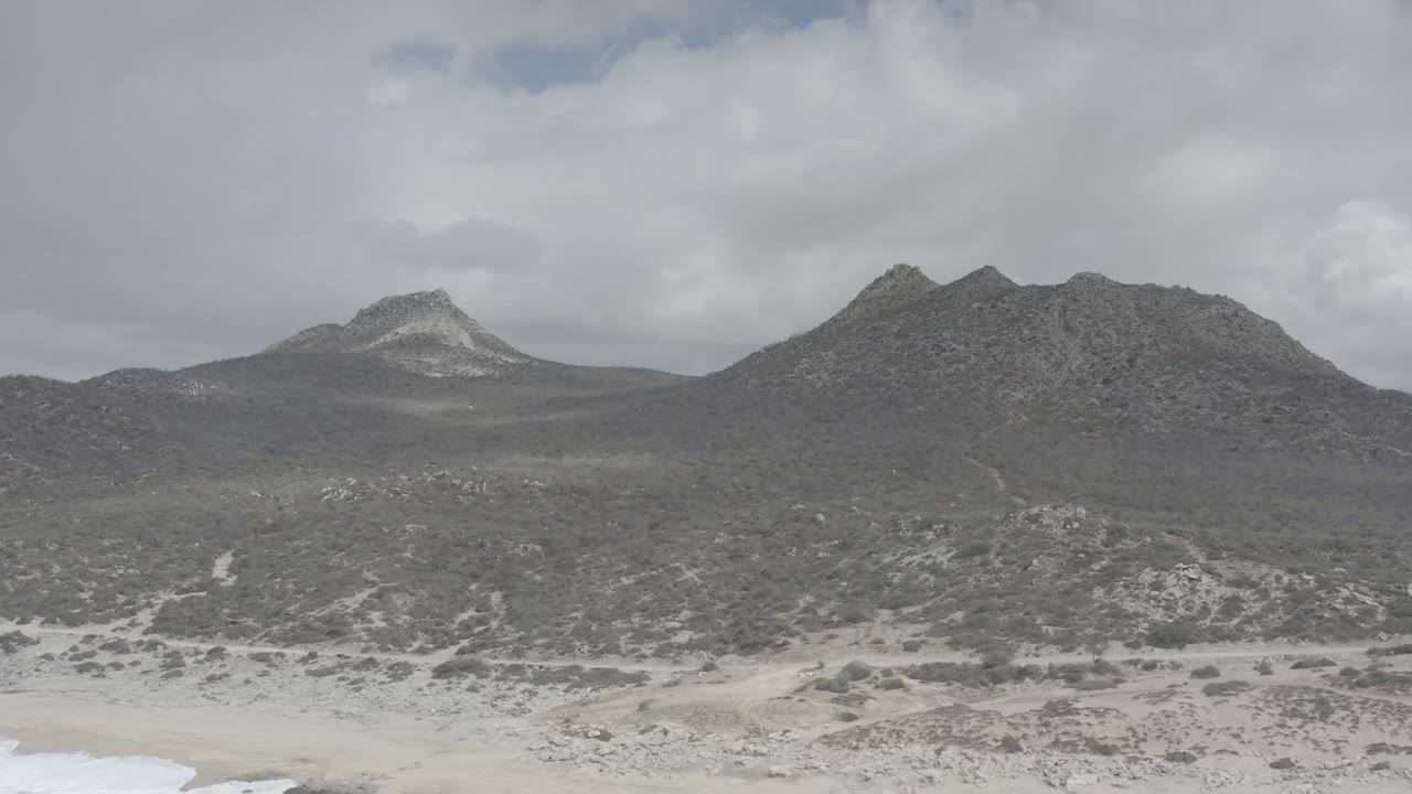 Wide elevated view in Los Cabos, Mexico, showing rocky hills across the horizon, gray-green brush in the midground, and a narrow band of pale beach at the bottom, no visible people.