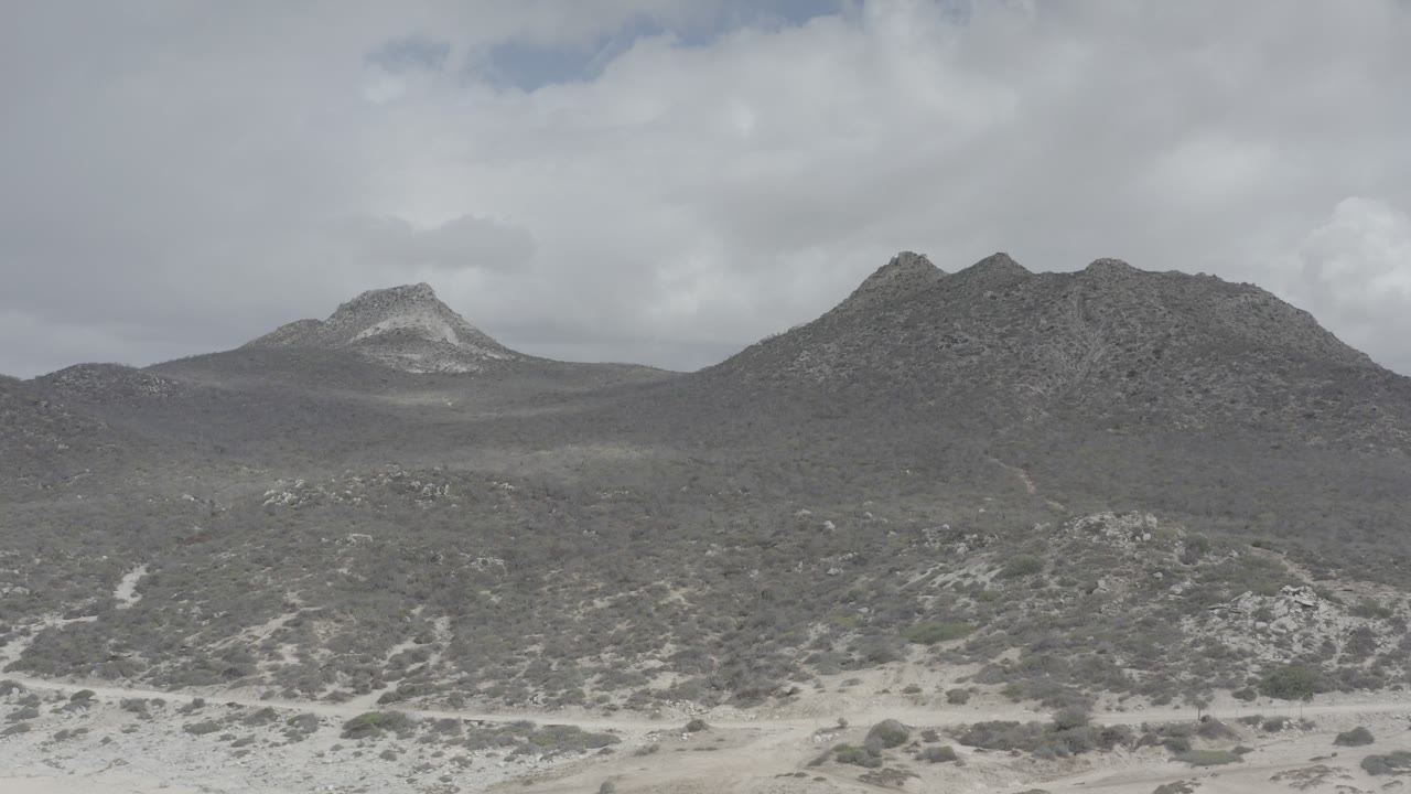 Wide elevated view of rocky mountains and sandy scrubland in Los Cabos, Mexico, under a gray cloudy sky with no visible people.