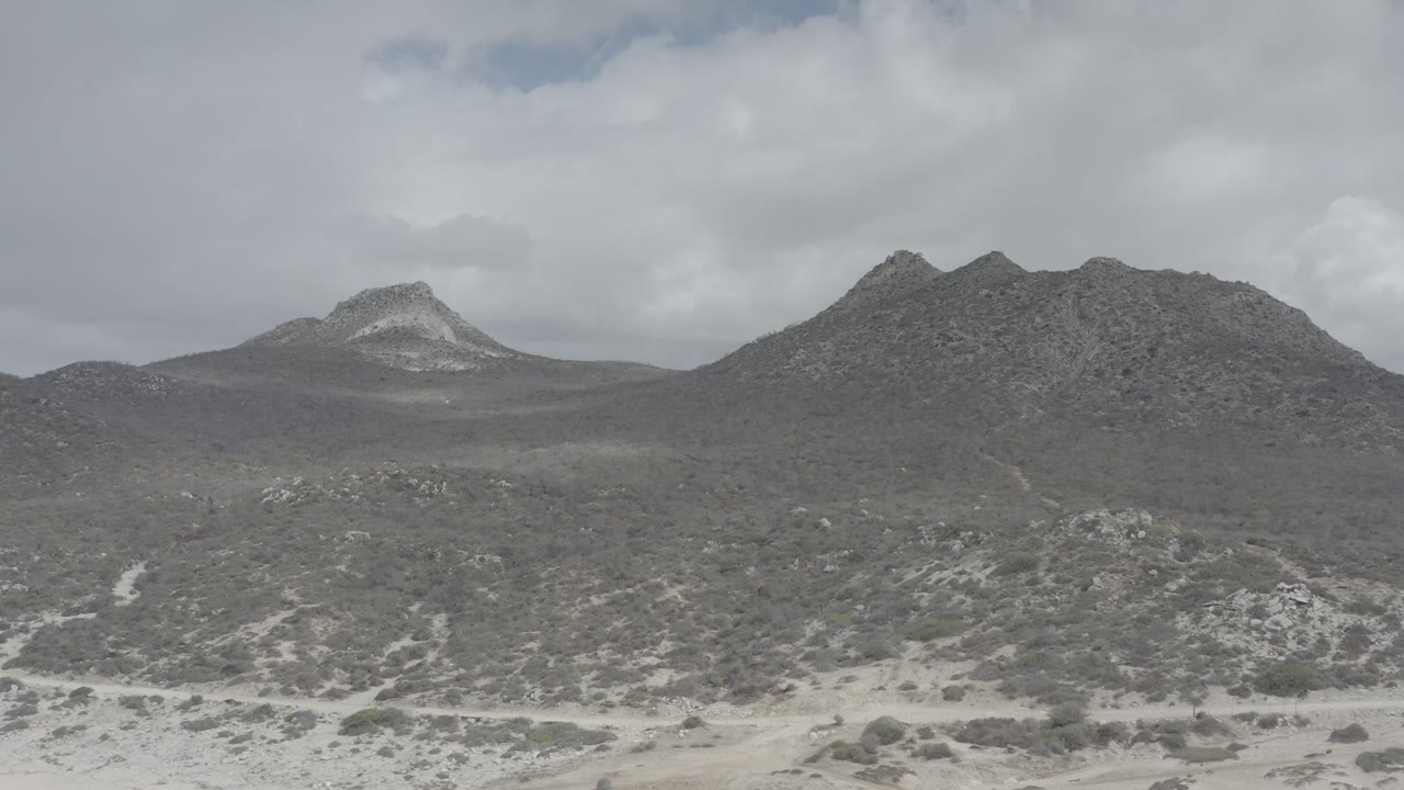 Wide view of the Los Cabos mountain ridge with pale sandy ground, low scrub, heavy gray clouds, and no visible people.