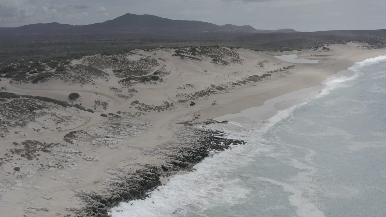 Aerial view of surf breaking along a sandy coast with dunes and hills inland.