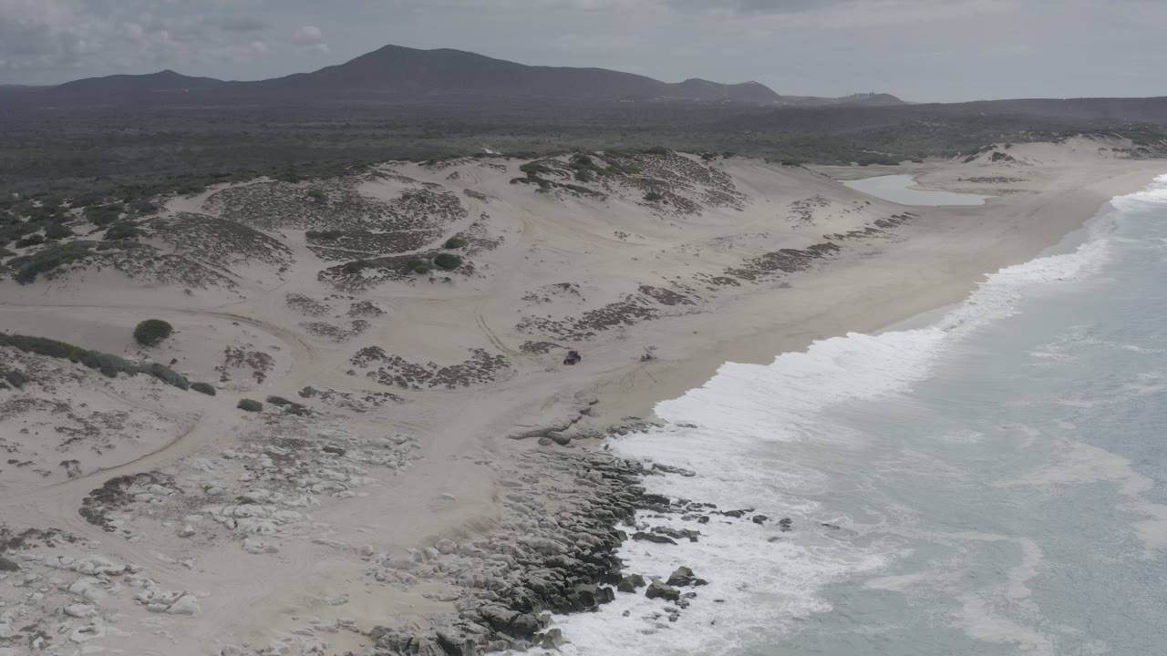 Aerial view in Los Cabos, Mexico of pale beach, foamy surf, scattered rock outcrops, dune vegetation, and dark hills on the horizon, with no visible people.