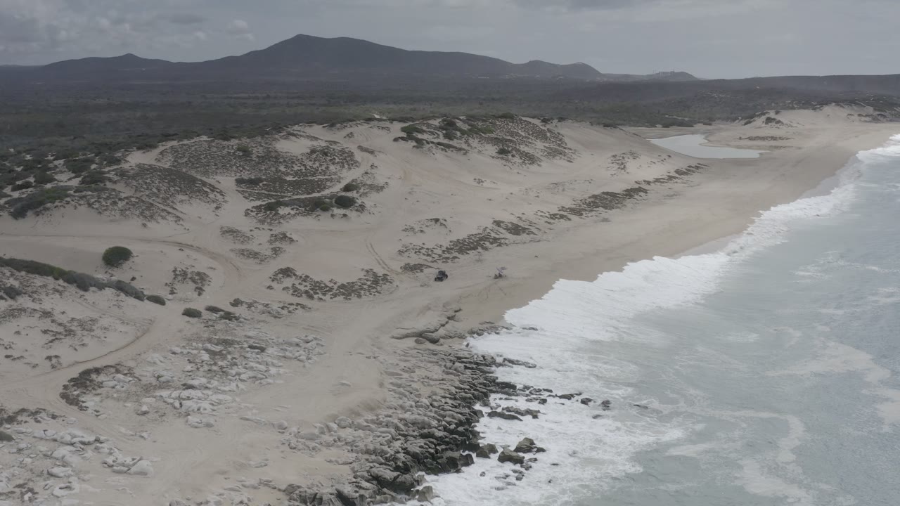 Los Cabos aerial frame showing white water rolling along the beach, rocky patches near the lower shore, dune scrub in the midground, and a dark hill line beyond, with no visible people.