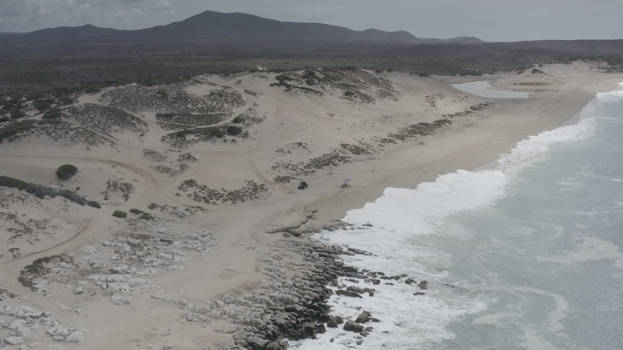 Aerial coastline with white surf, pale sand, dunes, and dark hills in the distance.
