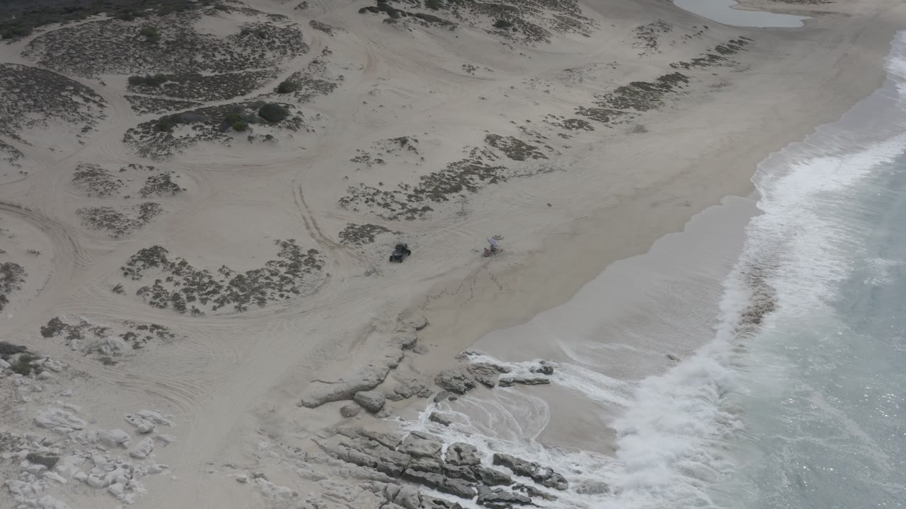 Aerial view over a Los Cabos, Mexico beach with pale sand, white surf along the right edge, low dune vegetation, rocks near the lower left, and no visible people.