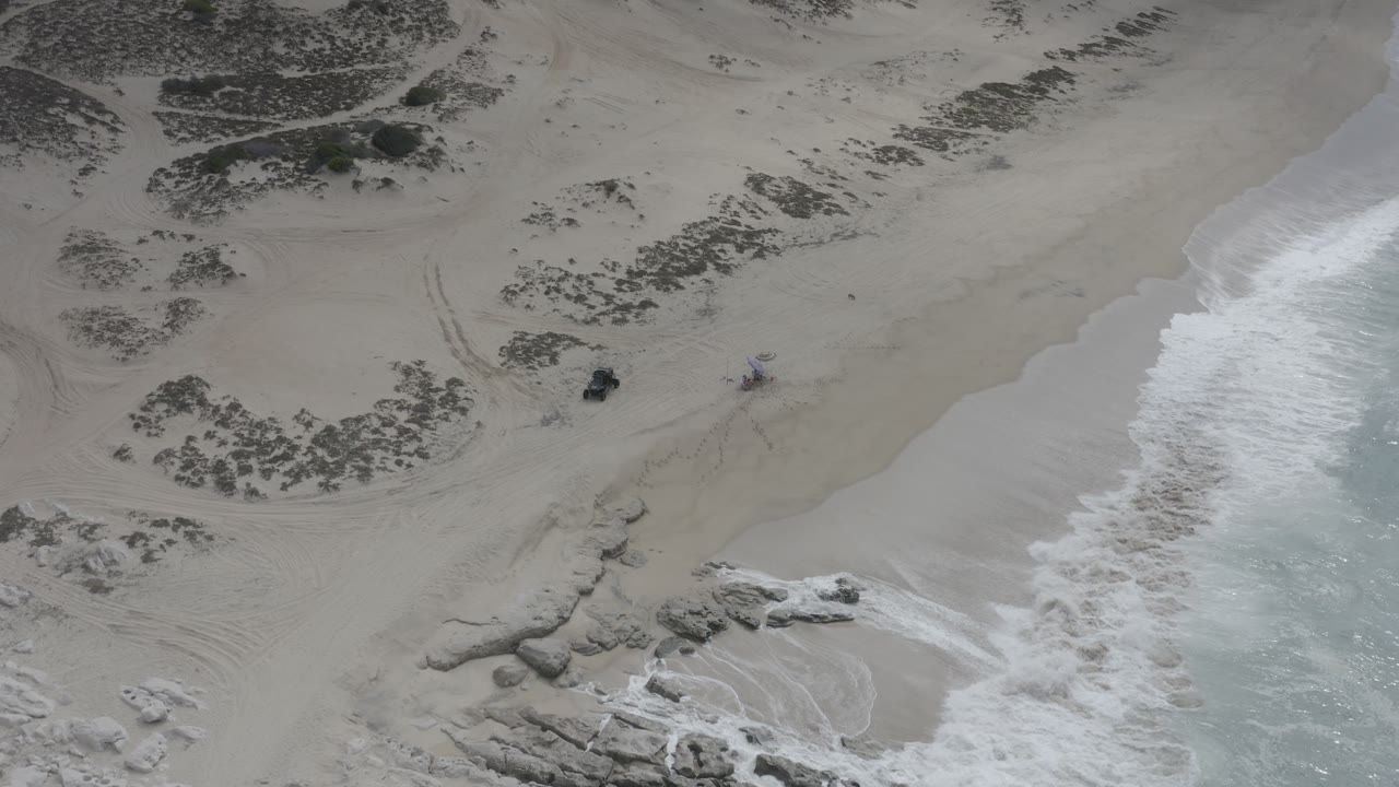 Overhead beach view with waves, tire tracks, scrub, and scattered rocks.